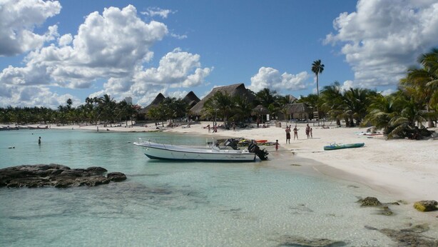 A beach with clear turquoise water, a boat docked near the shore, and thatched-roof huts in the background. - The Condo is in the gated Sirenis Resort community. (Akumal)
