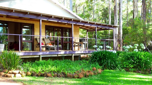 Una cabaña de madera con un porche cubierto, ventanas amplias y un jardín con rocas y plantas.