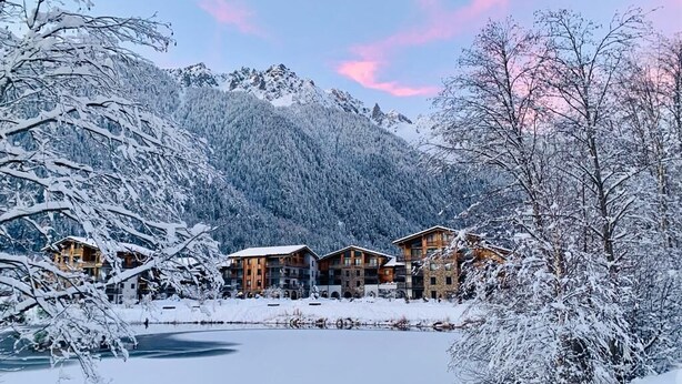 A snow-covered lodge with multiple wooden buildings, a frozen lake, and snow-covered trees. - 65m2 apartment, sleeps 6/7, lake and Mont Blanc view in ***** spa residence (Chamonix-Mont-Blanc)