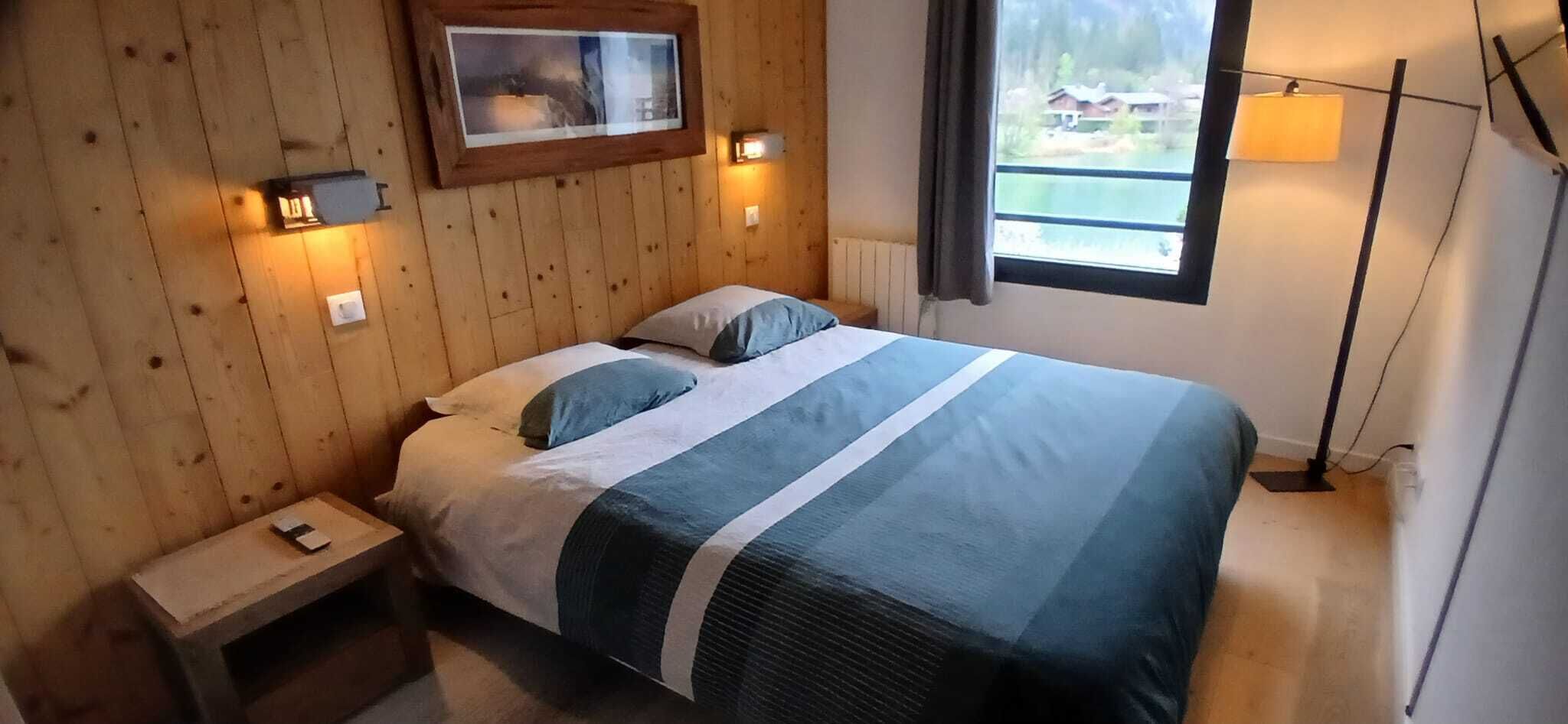 A wooden-paneled room with a bed, a nightstand, a framed picture on the wall, and a window with a view of a building and trees.