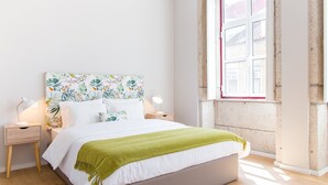 Ein ordentlich eingerichtetes Schlafzimmer mit einem Bett, zwei Nachttischen, einem Fenster mit Blick auf Gebäude und einer grünen Tagesdecke.