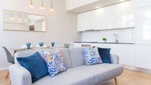 Ein modernes Wohnzimmer mit grauem Sofa, blauen Kissen mit Muster und einem Essbereich mit weißen Stühlen und blauem Geschirr.