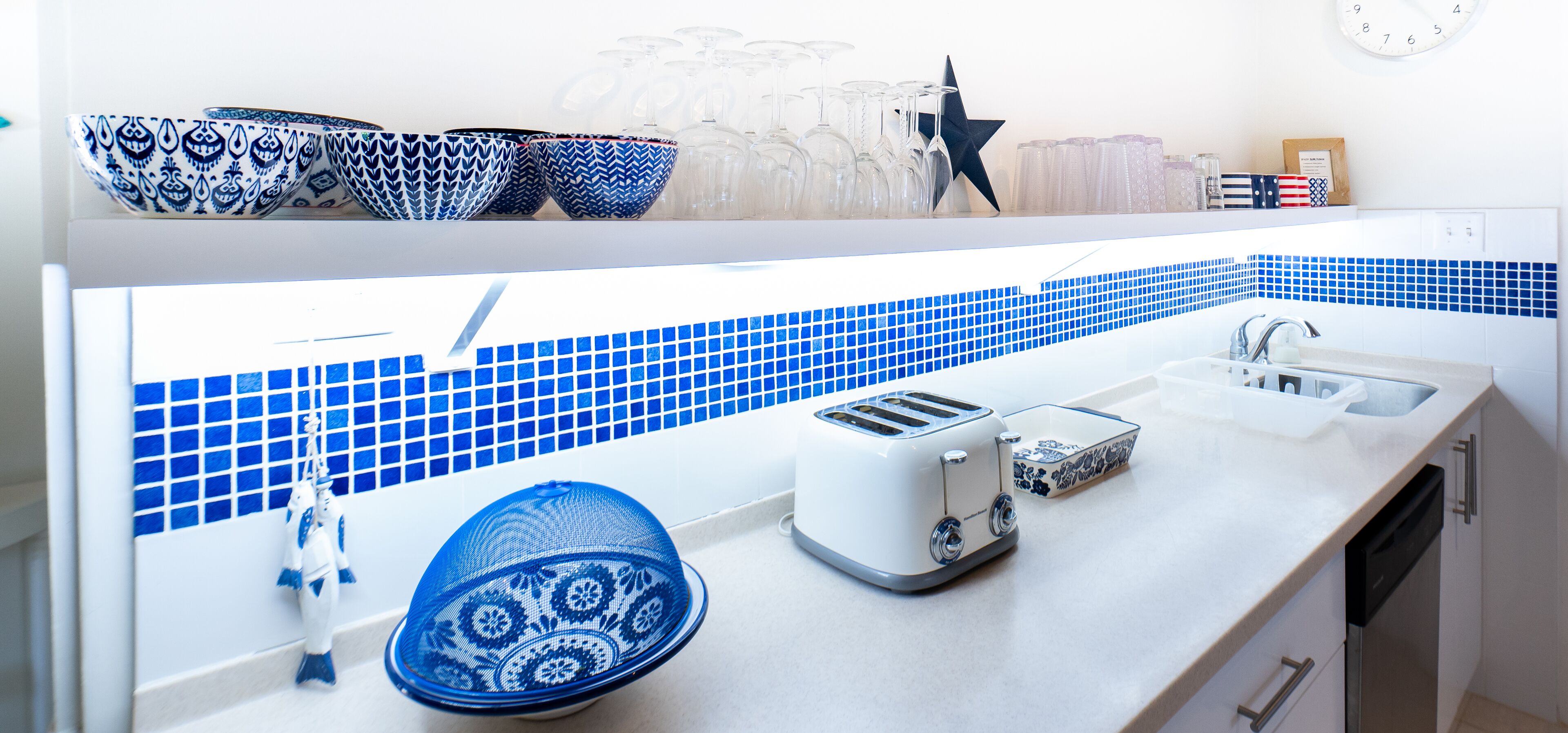 Una cocina con encimera blanca, salpicadero con azulejos azules, un tostador, un lavamanos y cuencos decorativos.