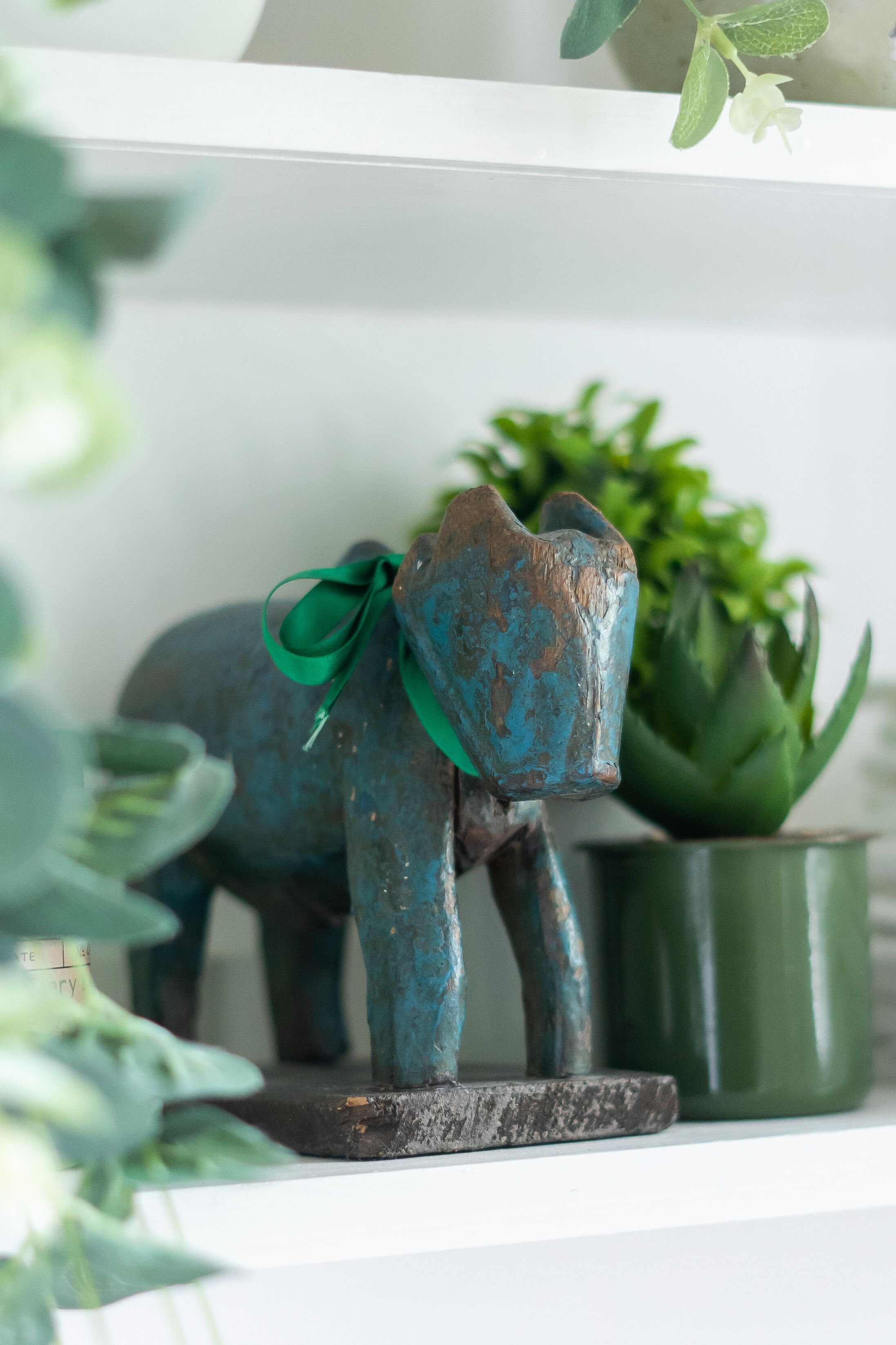 A rustic elephant sculpture with a green ribbon on a shelf, accompanied by a potted plant.