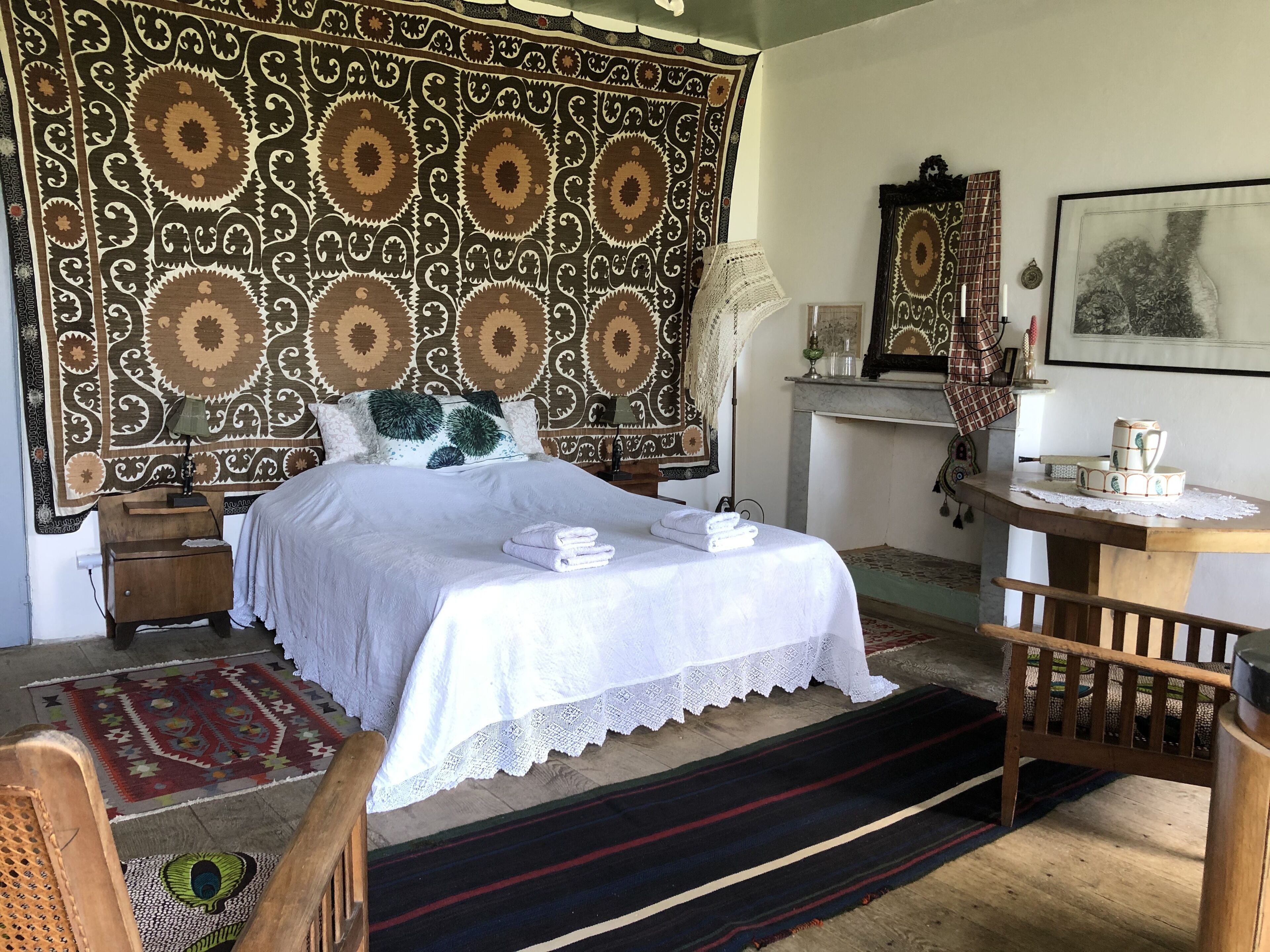 A neatly arranged bedroom with a large patterned tapestry, a bed with white linens, a wooden nightstand, a desk with a lamp, and a chair.