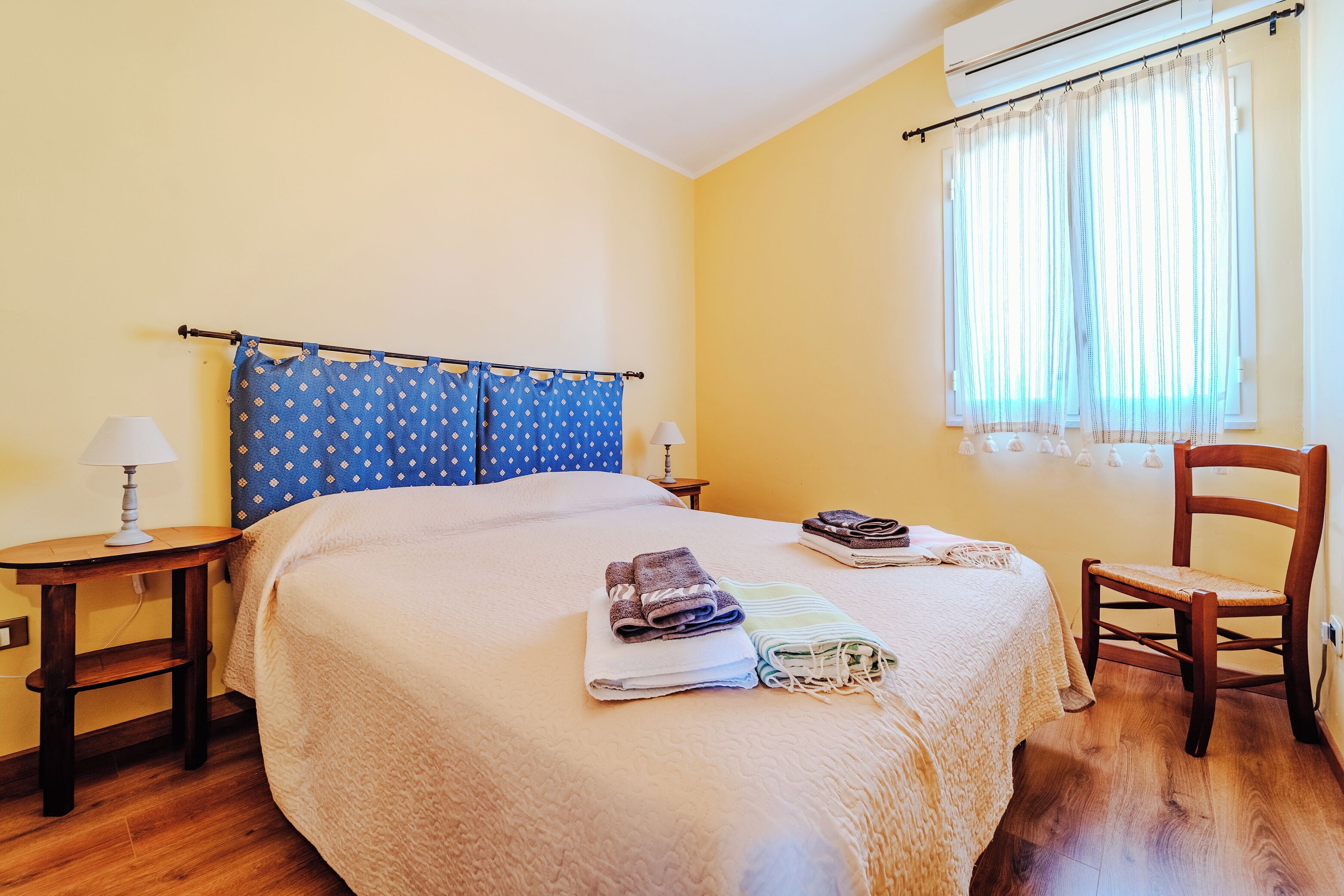 A neatly made bed with folded towels, a wooden chair, a small table with a lamp, and a window with curtains.