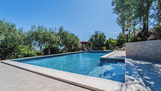 A rectangular swimming pool with a tiled deck, surrounded by trees and a stone wall. - Villa with exclusive swimming pool 5 minutes from the sea (Palermo)
