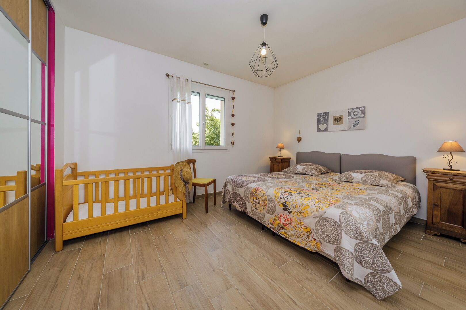 A bedroom with a wooden bed, a wooden chair, a bedside table with a lamp, a window with curtains, and a wall-mounted light fixture.