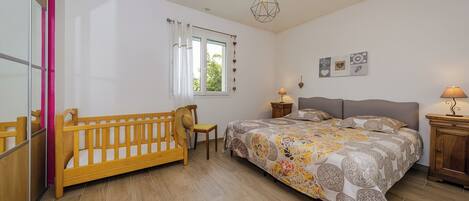A bedroom with a wooden bed, a wooden chair, a bedside table with a lamp, a window with curtains, and a wall-mounted light fixture.