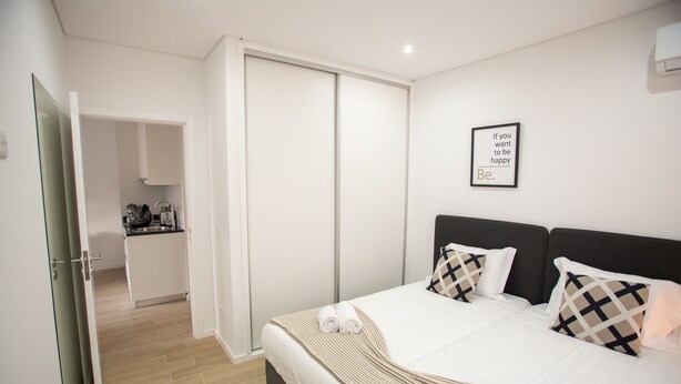 A hotel room with a bed, pillows, a wall-mounted picture, and a kitchen area visible through an open door.