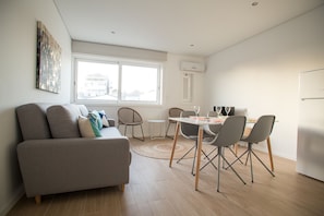 A modern living room with a sofa, dining table, and chairs. There is a window providing natural light and a view of buildings outside. - Camões 691 Design Apartments by Porto City Hosts (Porto)