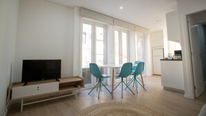 A modern living room with a flat-screen TV, a dining table with chairs, and a kitchen area visible through the window.
