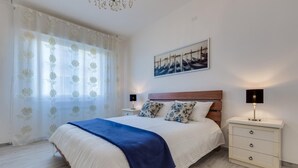 Un dormitorio con una cama grande, un cabecero de madera, dos mesitas de noche con lámparas, una ventana con cortinas traslúcidas y un cuadro en la pared.