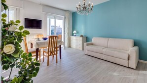 Una sala de estar moderna con un sofá blanco, una mesa de comedor con sillas, un televisor montado en la pared y una araña.