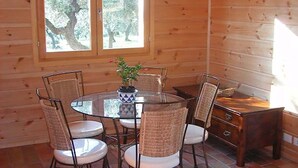 L’intérieur d’un chalet en bois présente une table ronde en verre entourée de chaises en osier, une commode en bois et une fenêtre donnant sur la forêt.