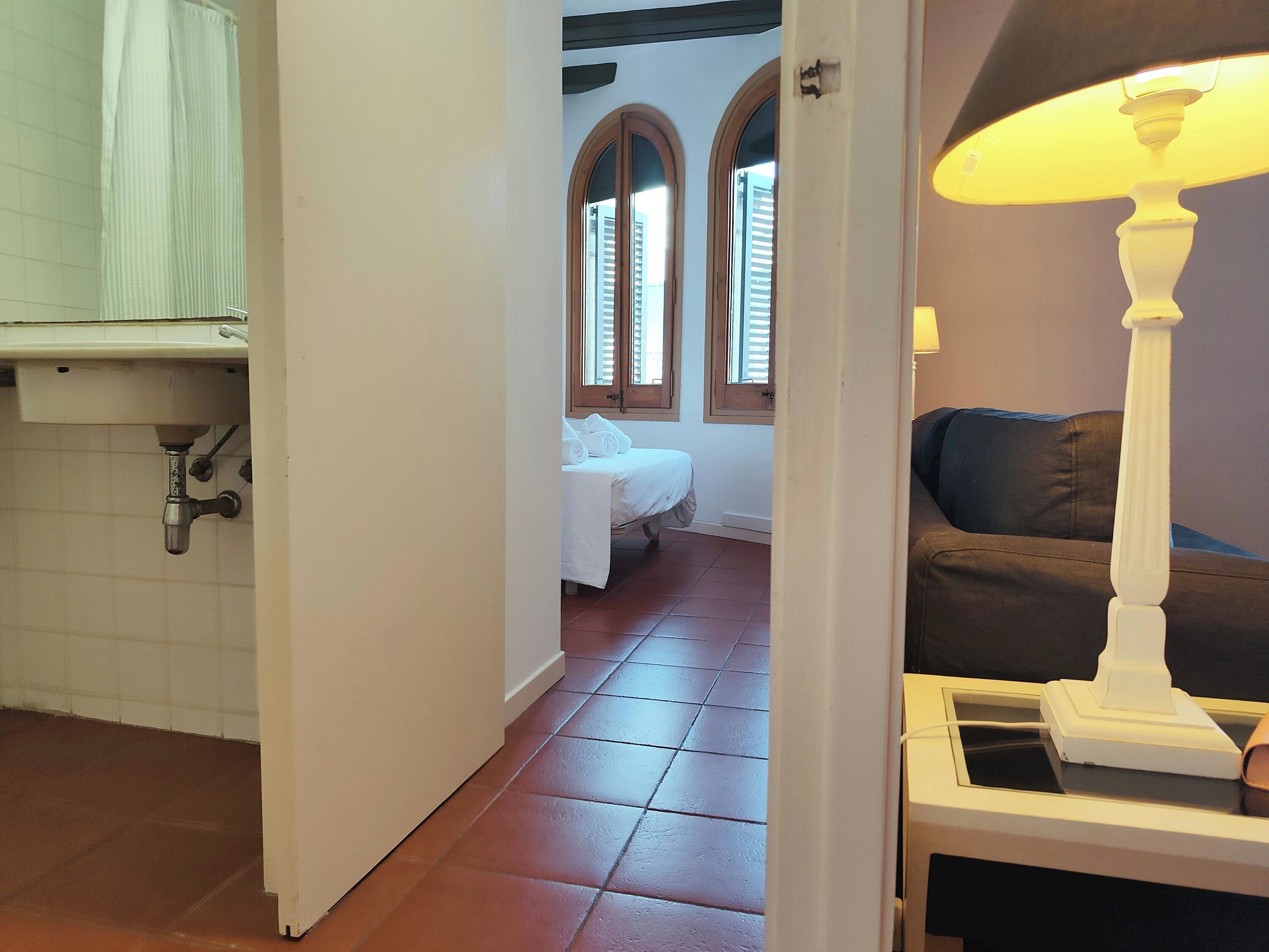 Una habitación con una cama, una ventana con contraventanas, una lámpara y un piso de baldosas.