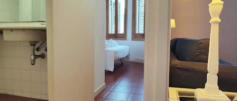Una habitación con una cama, una ventana con contraventanas, una lámpara y un piso de baldosas.