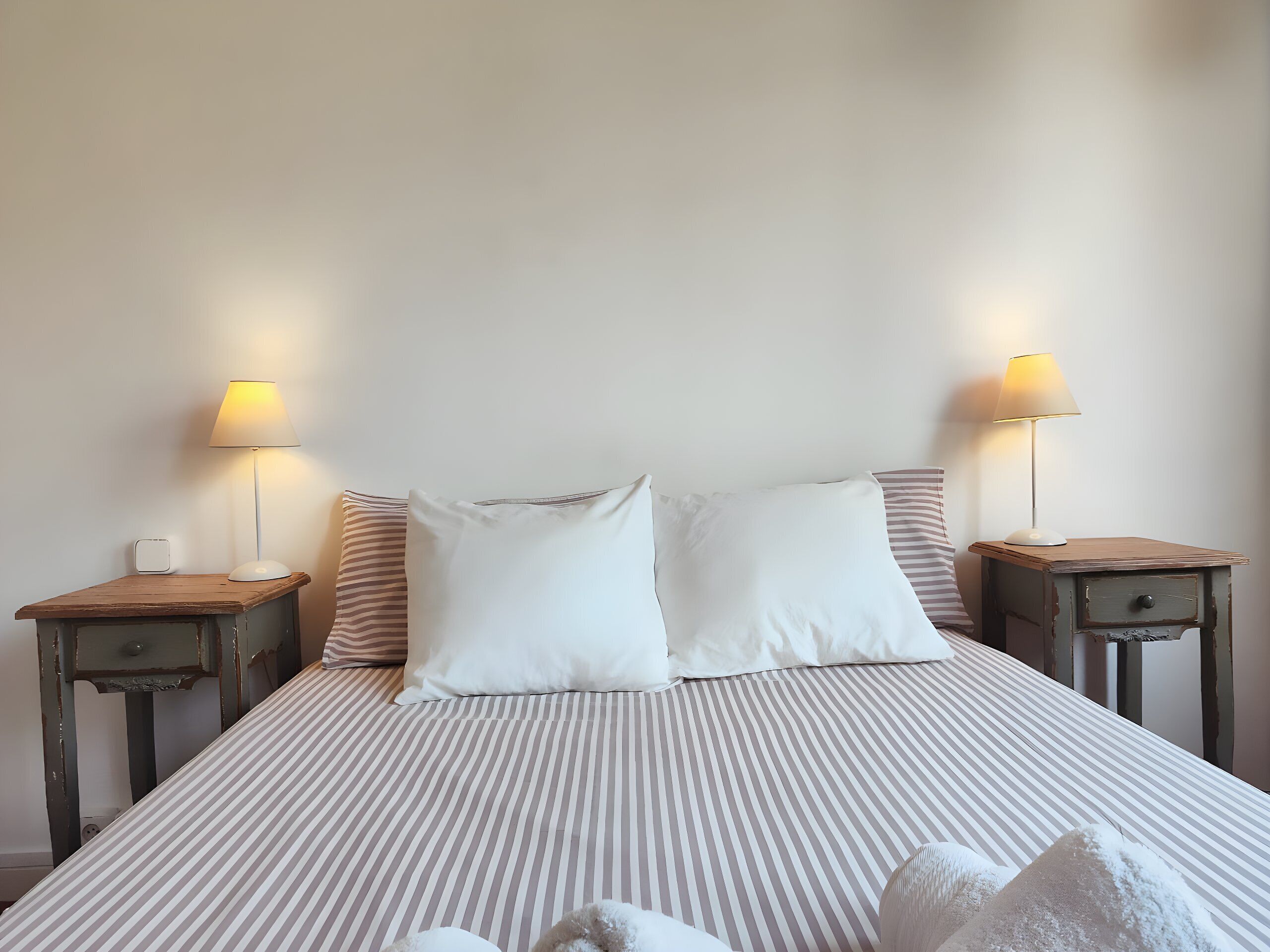A bed with striped bedding, two bedside lamps, and a wooden nightstand with a drawer.