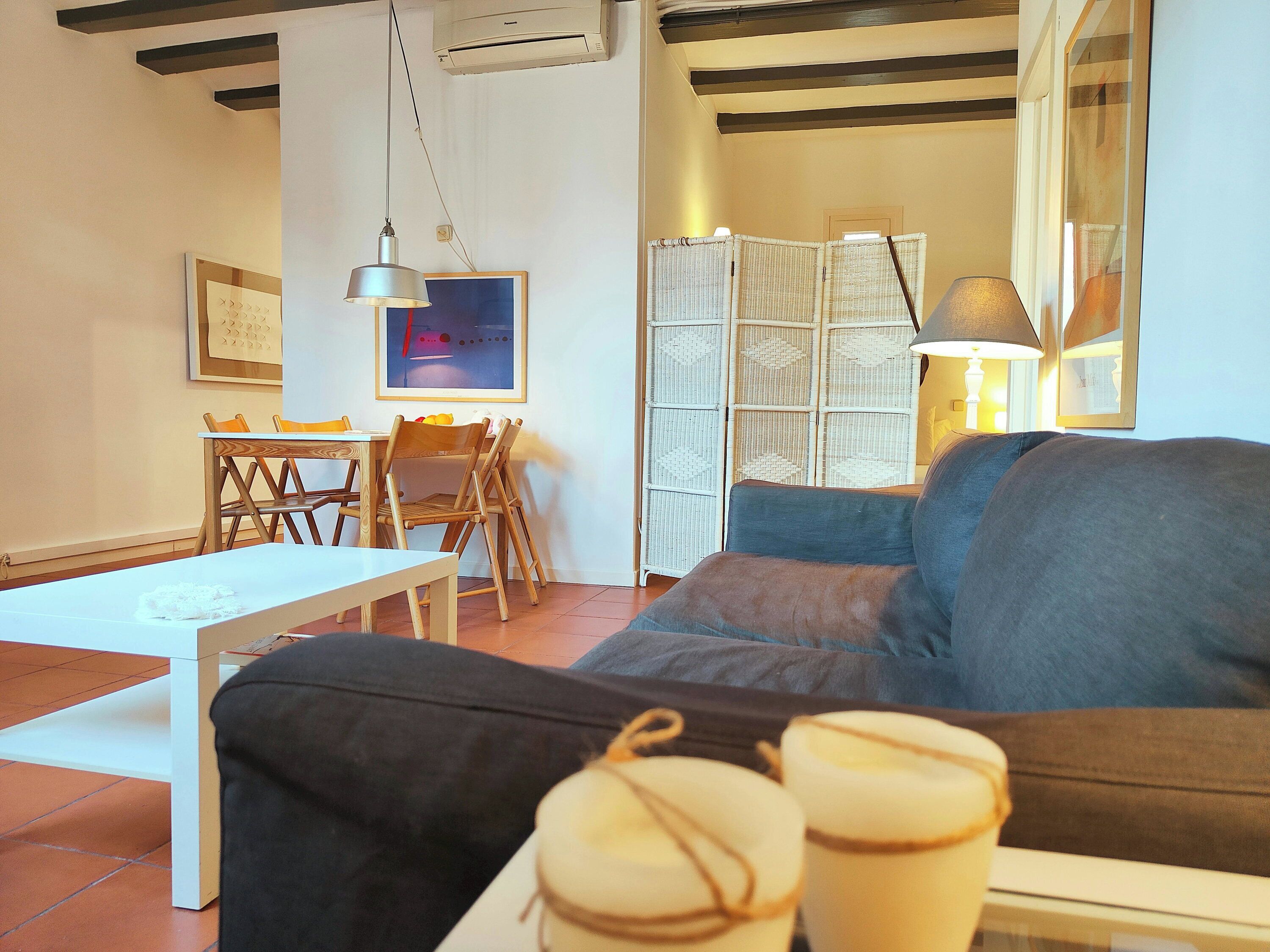 Ein Wohnzimmer mit einer Couch, einem Tisch und Stühlen. Eine Lampe, eine Klimaanlage und ein gerahmtes Bild sind an der Wand zu sehen.