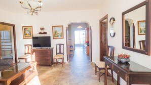Amplia sala de estar con televisión, muebles de madera, una araña y un pasillo que conduce a otra habitación.