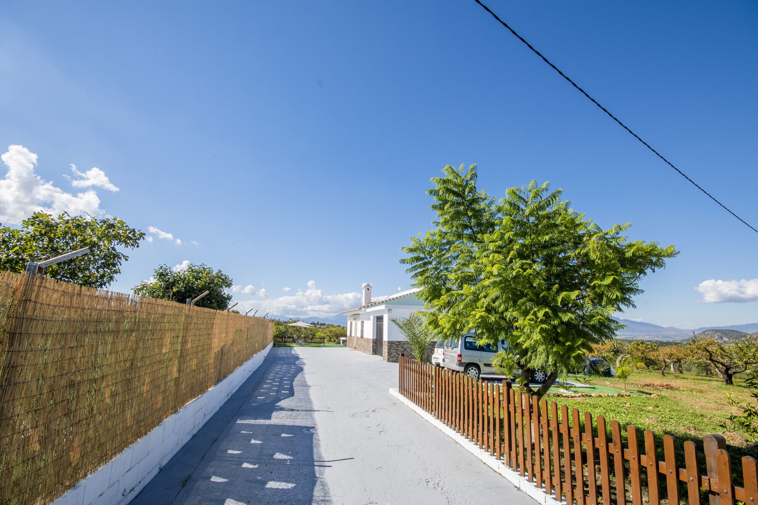 Eine schmale Straße, begrenzt von einem Holzzaun und einer weißen Mauer, die zu einem Haus mit Schornstein führt.