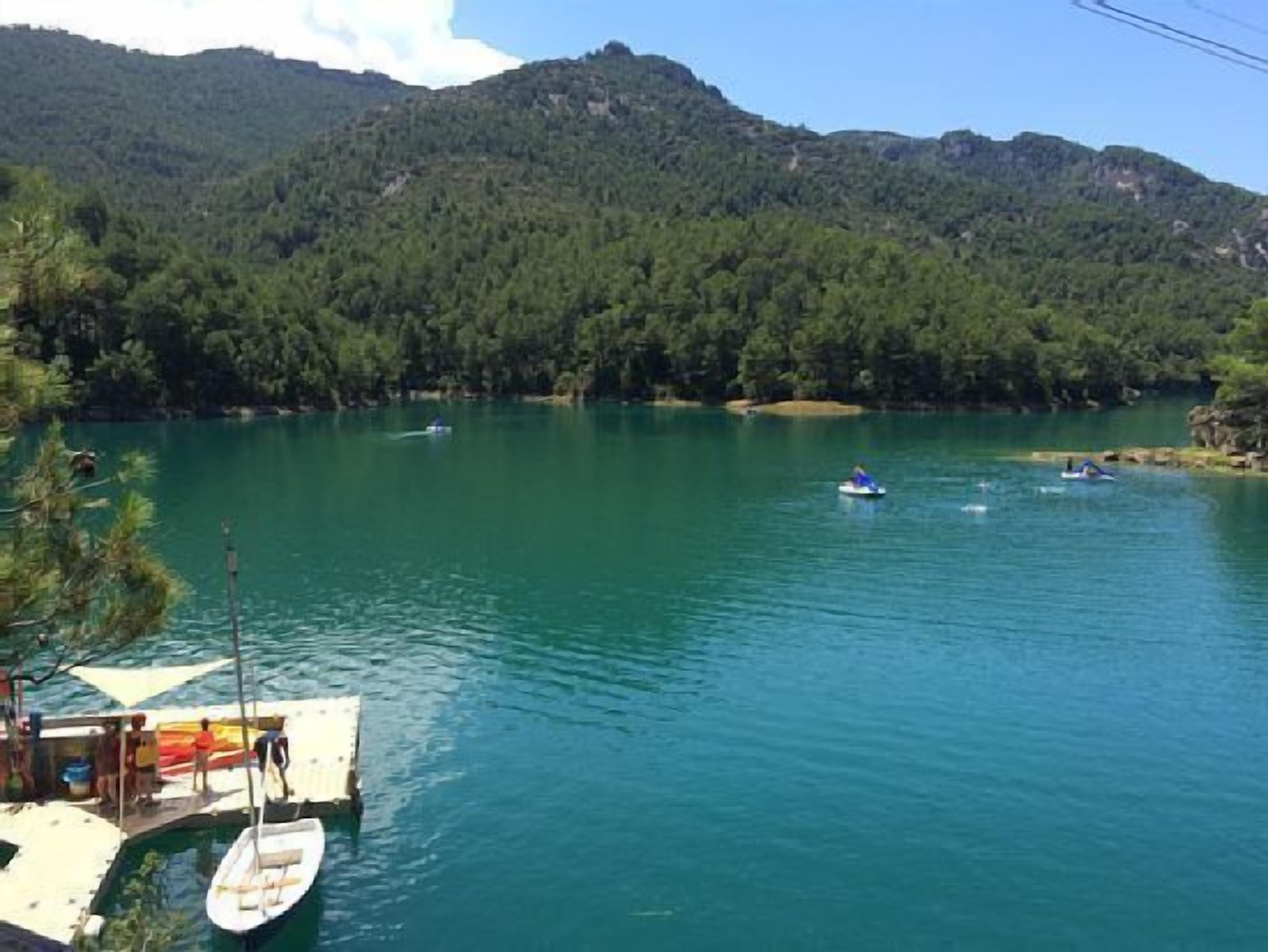 A calm lake surrounded by forested hills, with boats on the water and a dock with a boat tied to it.