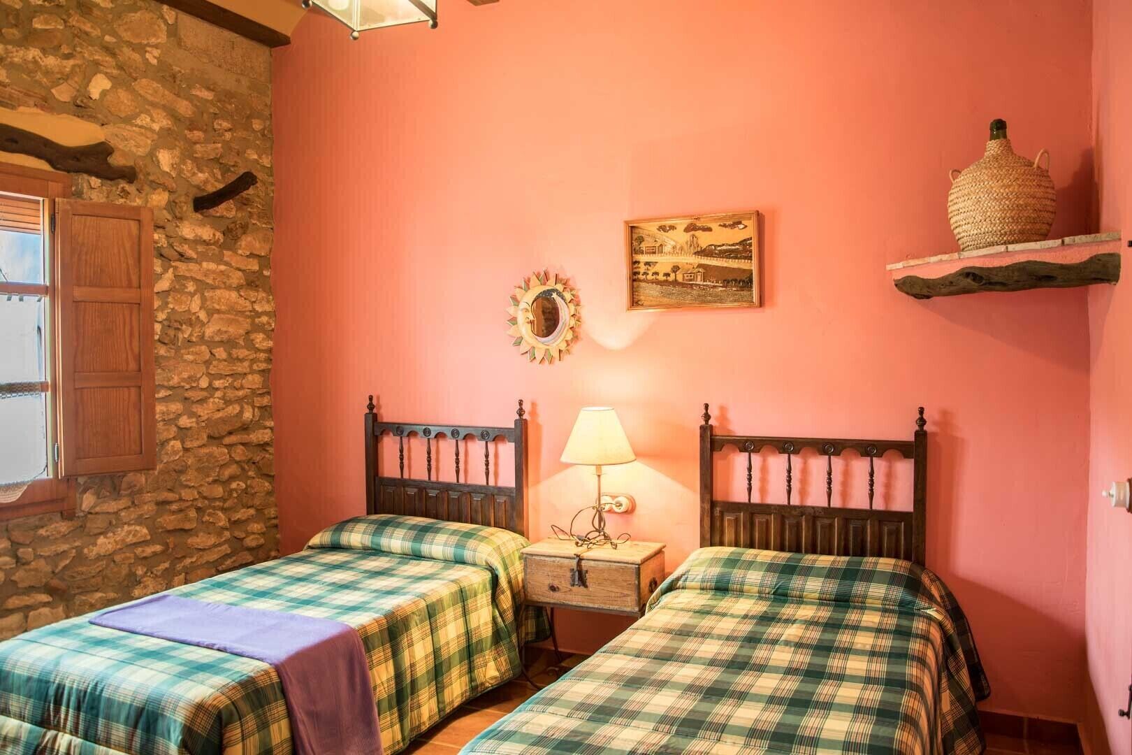 A room with two beds, a stone wall, a wooden ceiling, and a small table with a lamp.