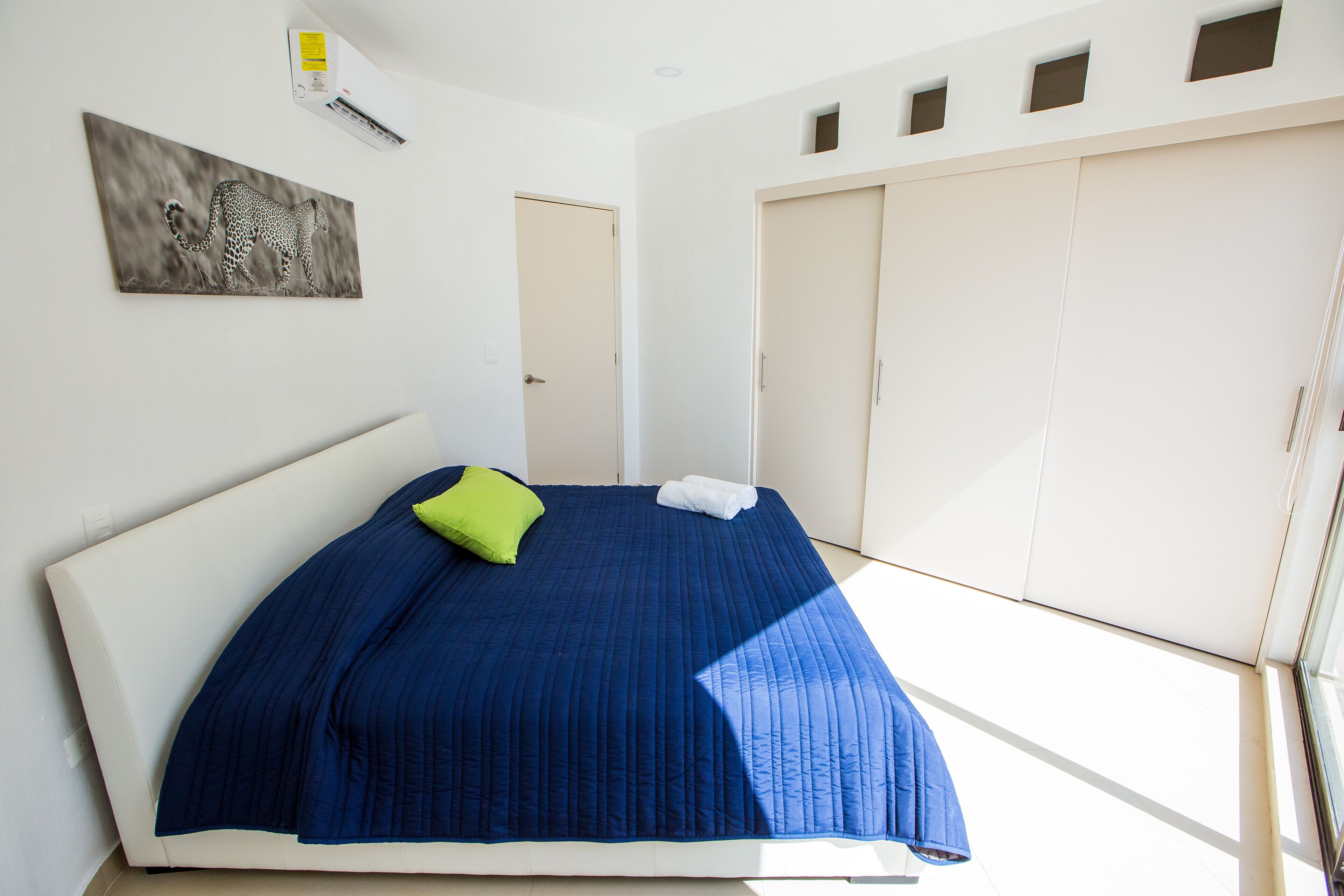 A bedroom with a bed, a green pillow, a blue bedspread, and a painting on the wall.