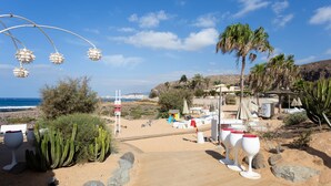 A beachside resort with a wooden deck, palm trees, cacti, and beach chairs.