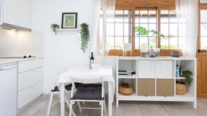 A modern kitchen with white cabinetry, a dining table with chairs, a window with sheer curtains, and a wooden bench with baskets.