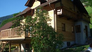 A two-story wooden house with a balcony, surrounded by greenery and a mountain in the background.