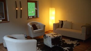 A living room with a white sofa, a white armchair, a small round table, a cowhide rug, and a window with a view of greenery.
