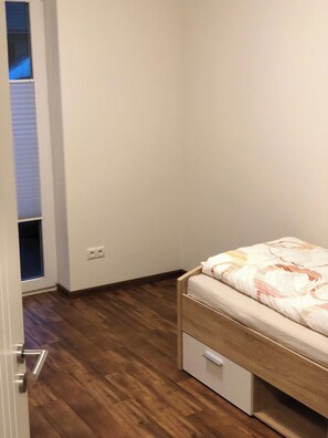 Un dormitorio con una cama de madera, un cajón pequeño blanco y una puerta que da a un balcón.
