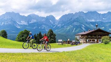 Ein Mountainbike-Weg mit zwei Radfahrern, ein Holzhaus mit Balkon und ein Berg im Hintergrund.
