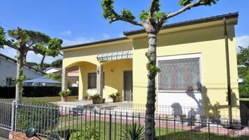 Une maison jaune avec une cour clôturée, un scooter garé et un arbre au premier plan.