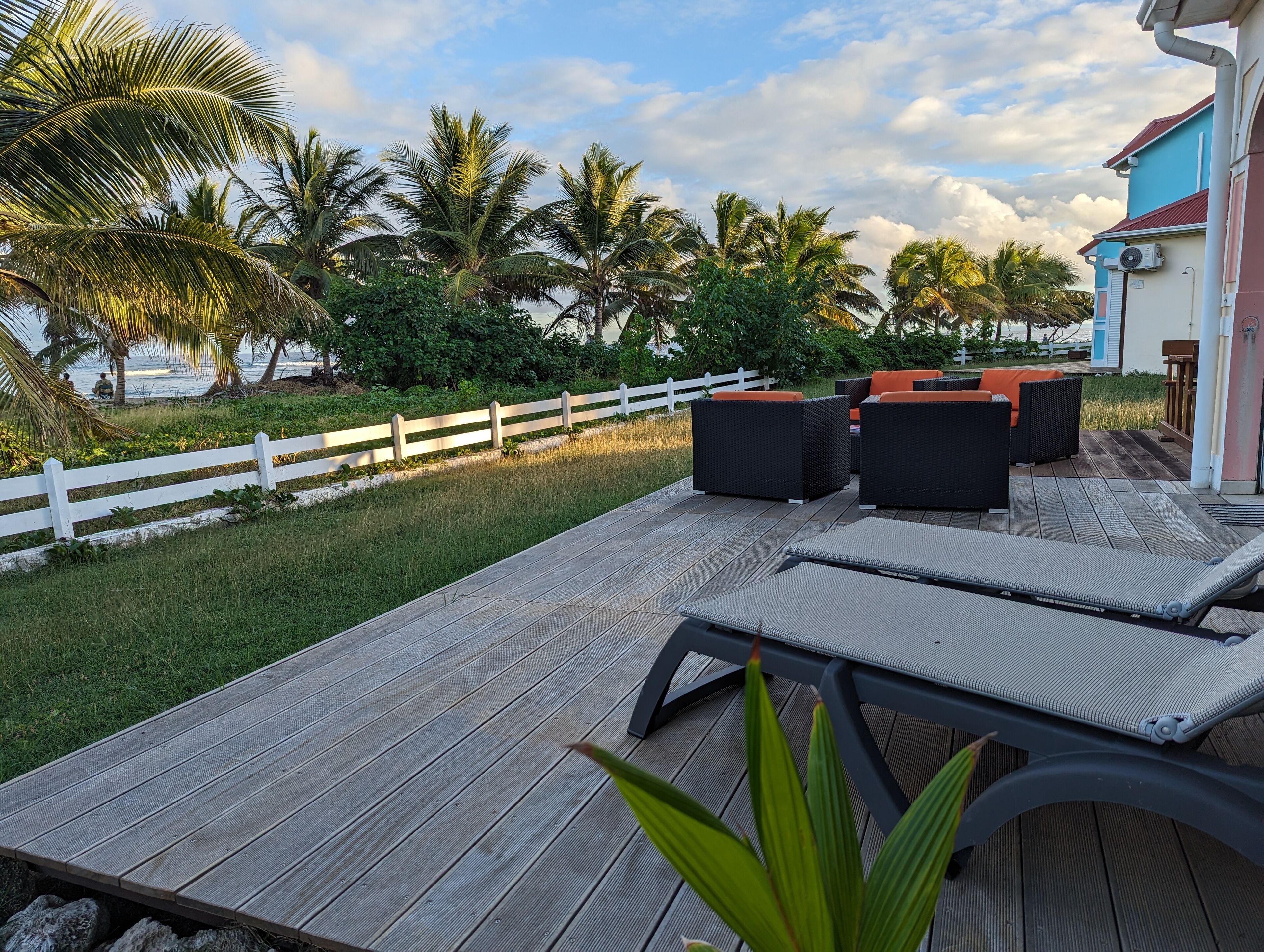A wooden deck with outdoor furniture, a white fence, and palm trees.