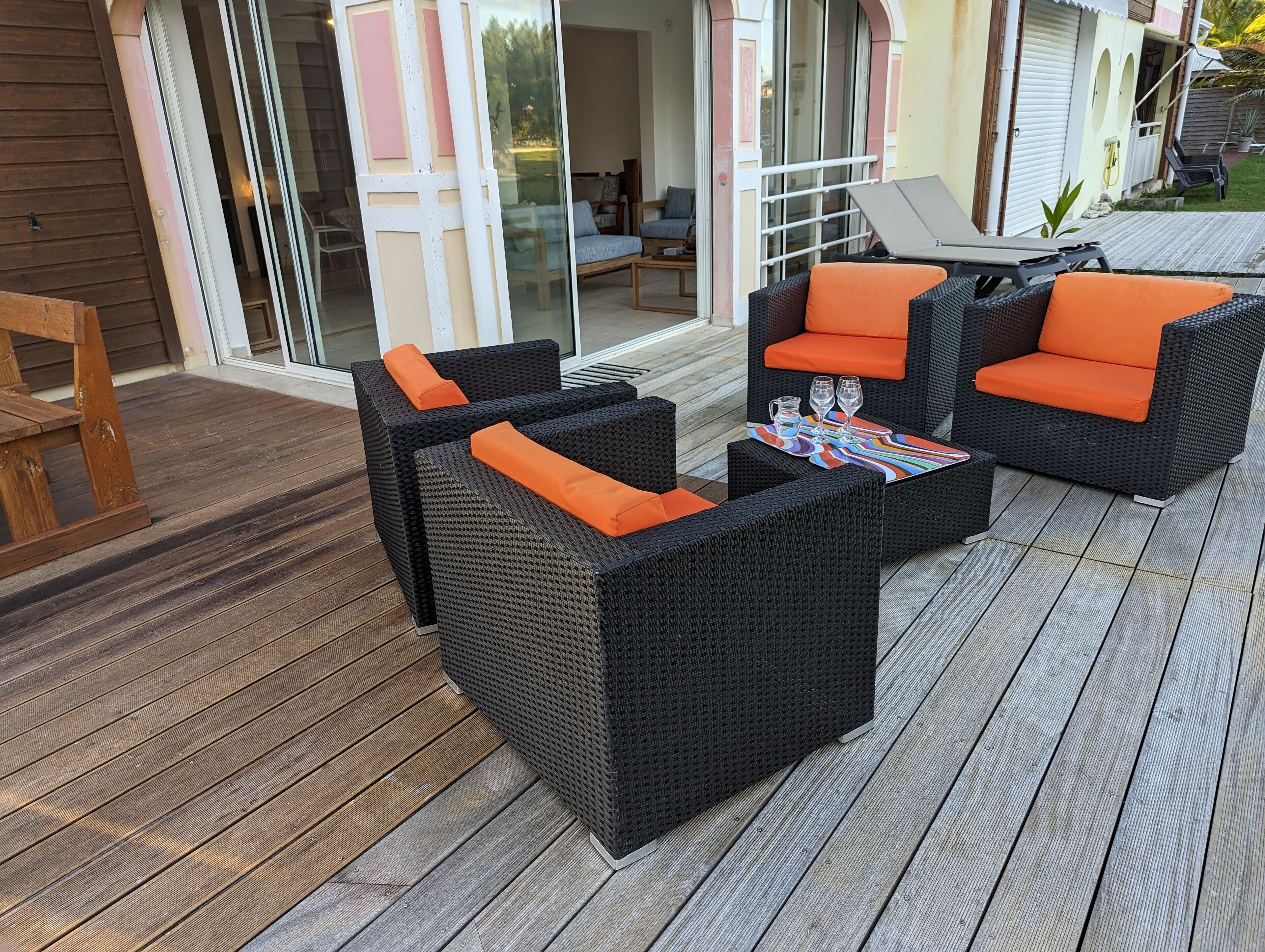 Une terrasse extérieure aménagée avec des meubles en osier, dotée de coussins orange, une petite table avec un set de table coloré et une porte vitrée donnant sur un espace abrité.