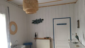 A small, cozy room with a white door, a blue chair, a round mirror, a wooden shelf with decorative items, and a hanging light fixture. - In Dinard, house with garden close to the sea - Gîte "Aod Gwer-Vein (Dinard)