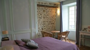Une chambre avec un mur en pierre, un lit recouvert d’une couverture violette, une chaise et une petite table.