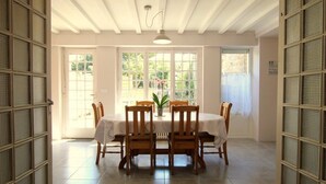 Un coin repas avec une table ronde, des chaises en bois, un ventilateur de plafond et de grandes fenêtres donnant sur un paysage verdoyant.