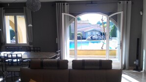 A living room with a sofa, a dining table with chairs, and a view of a swimming pool and outdoor area.