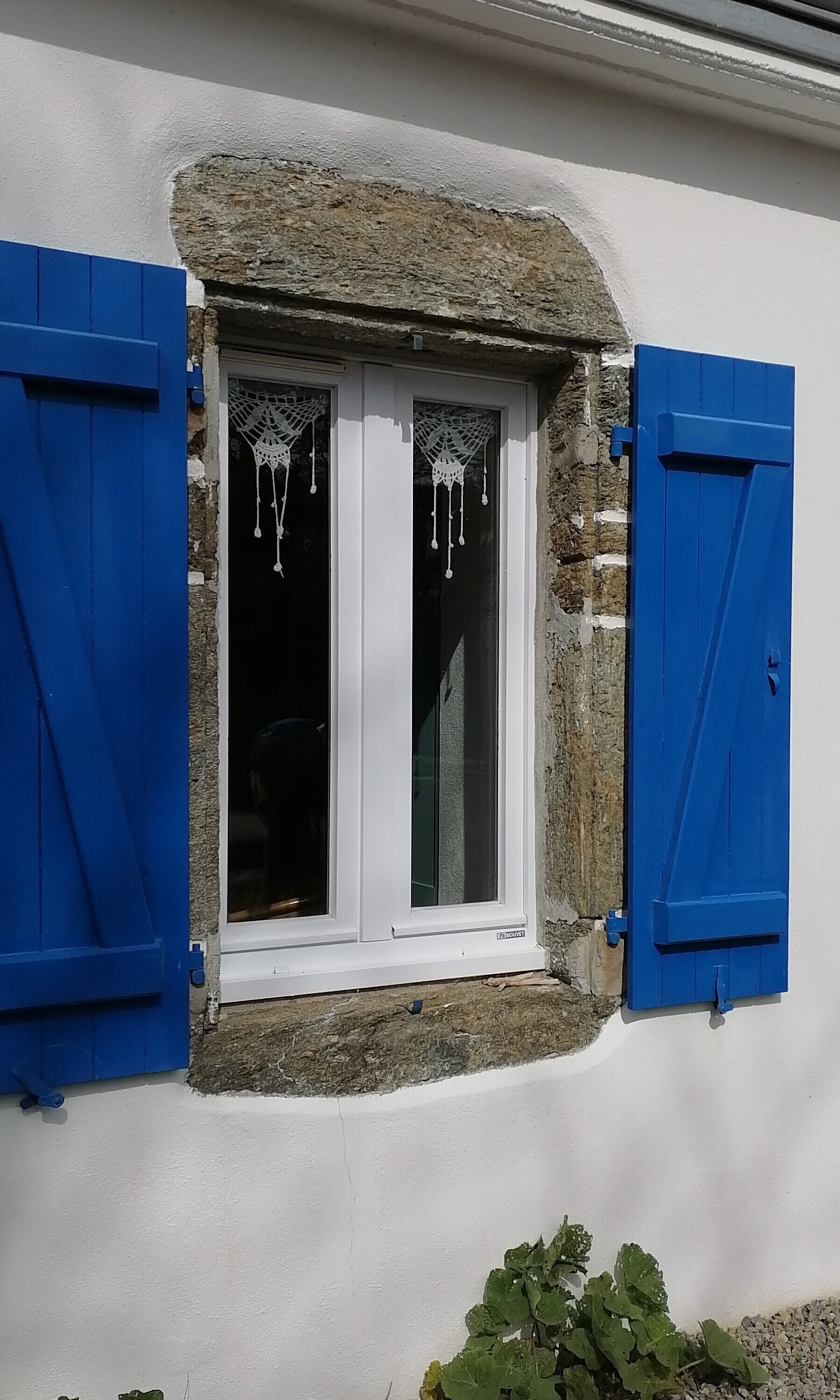 Una finestra con cornice bianca e persiane blu, incastonata in un muro in pietra con rivestimento bianco.