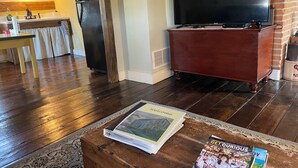 Un salón con piso de madera, un televisor sobre un mueble bajo, un libro sobre una mesa de centro y una cocina al fondo.