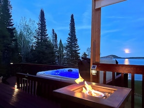 A hot tub on a deck with a view of a body of water and trees at dusk.