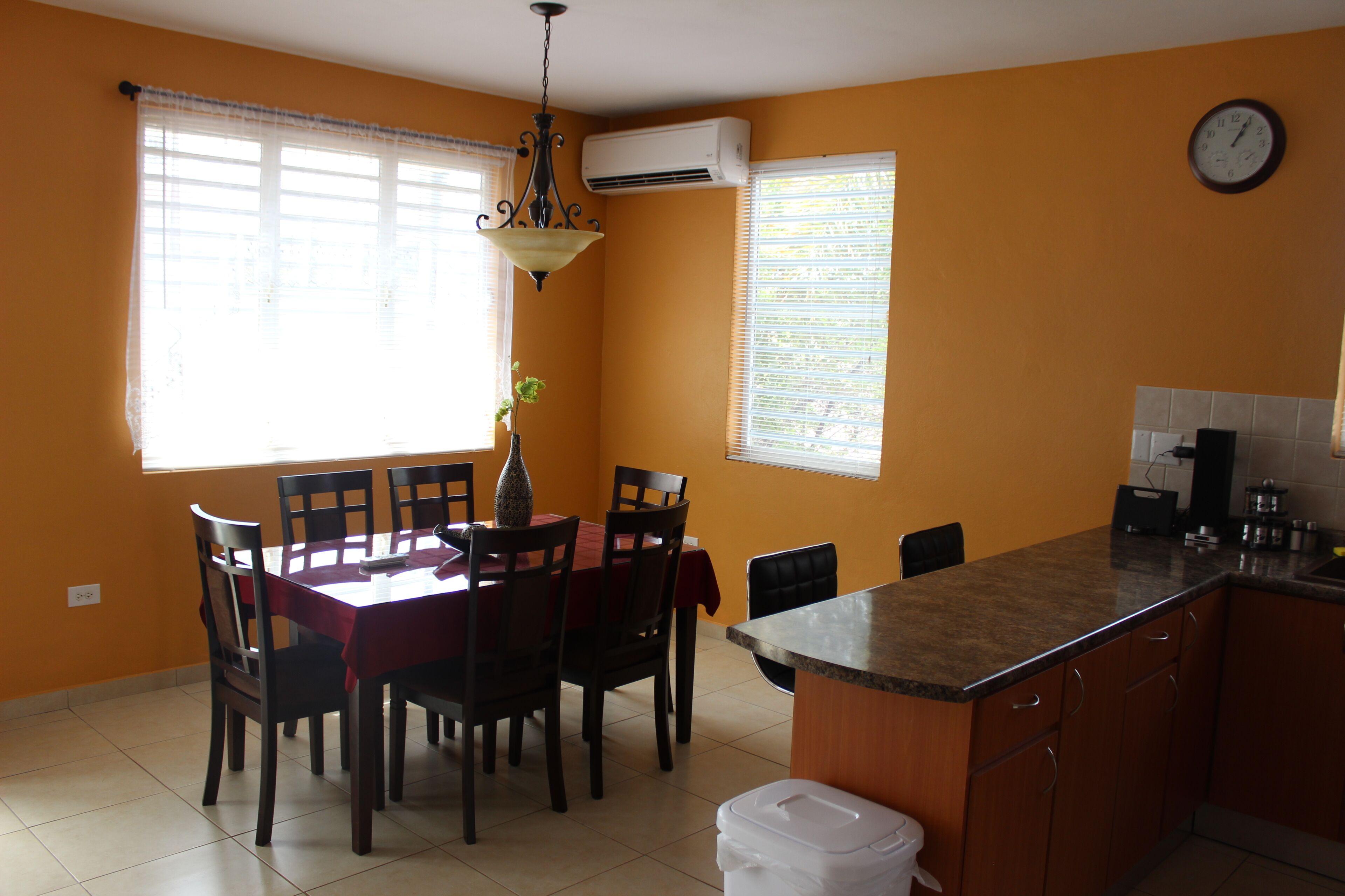 A dining area with a table and chairs, an air conditioning unit, a clock, and a kitchen counter.