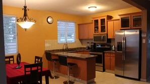 Una cocina con isla central, armarios de madera, un refrigerador de acero inoxidable y un comedor con mantel y sillas rojos.