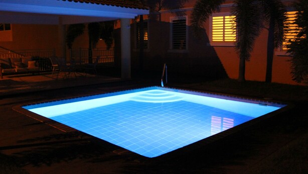 Una piscina con un diseño de cuadrícula en el piso, rodeada por un porche cubierto con sillas y barandilla.