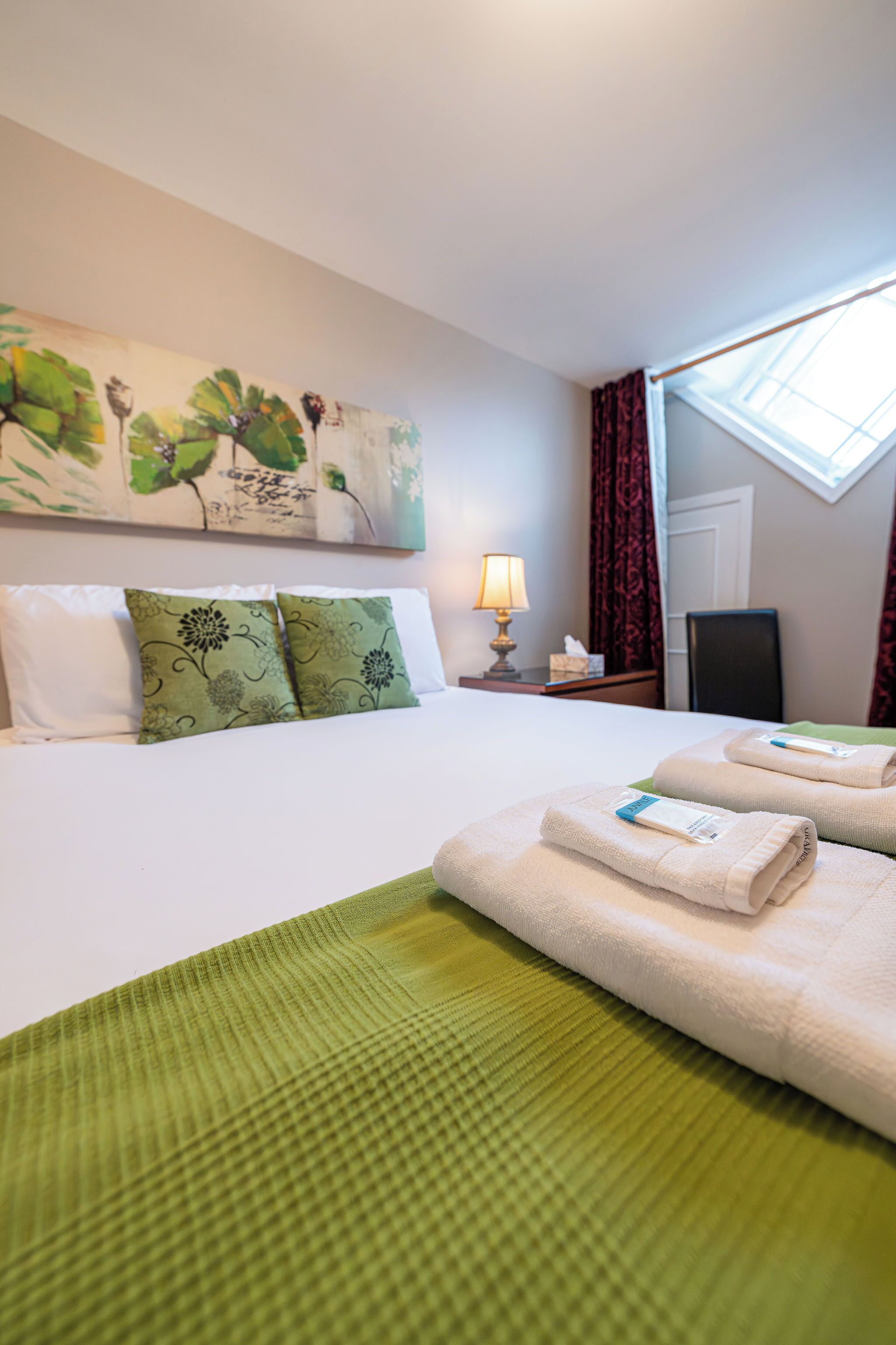 A hotel room with a bed, green and white pillows, a painting on the wall, a lamp, and a window with curtains.