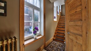Un escalier en bois, avec un tapis à motifs au bas, une fenêtre avec un pot de fleurs et un radiateur sur le côté gauche.