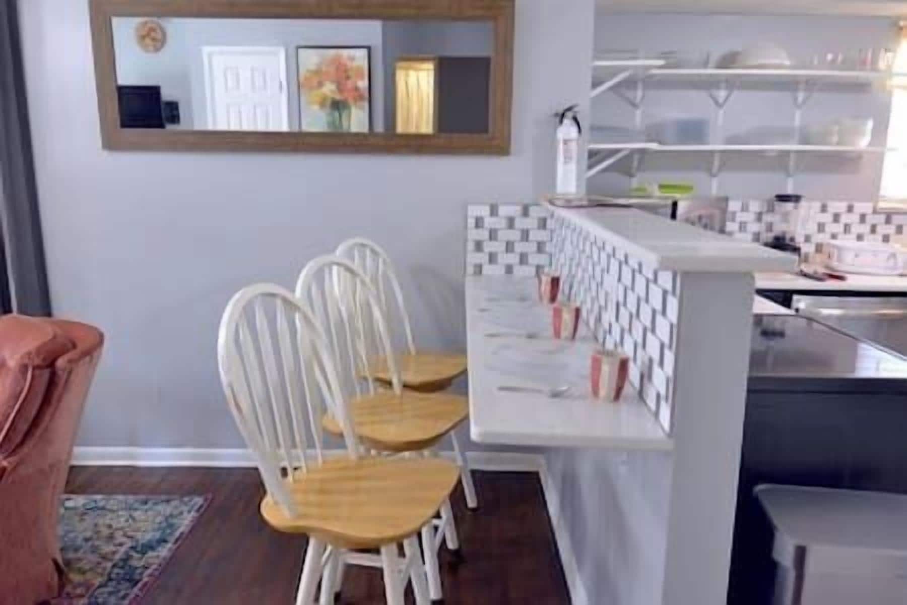 Une cuisine avec un plan de travail blanc, deux chaises blanches et un miroir au mur.